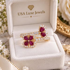 May include: A gold ring featuring two flower-shaped motifs set with pear-cut deep pink stones and surrounded by small clear stones. The ring is displayed in a cream-colored velvet box with the text USA Luxe Jewels printed on the inner lid.