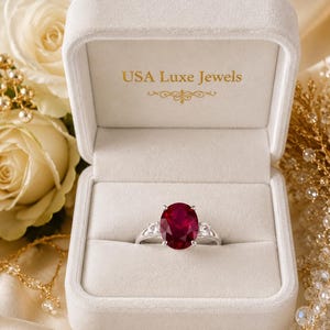 Puede incluir: Un anillo de plata con una gran piedra central de rubí rojo intenso de corte ovalado, flanqueada por dos piedras transparentes más pequeñas de corte redondo. El anillo se presenta en un joyero de terciopelo crema con el texto USA Luxe Jewels en oro.