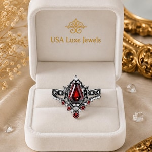 May include: A black metal ring featuring a central coffin-shaped red gemstone, accented with smaller round red stones, clear crystals, and crescent moon motifs. The ring is displayed inside a white velvet jewelry box labeled USA Luxe Jewels.
