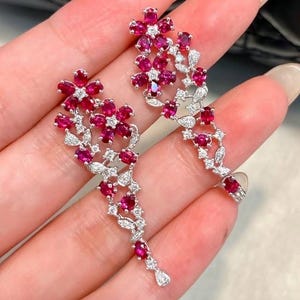 Puede incluir: Un par de pendientes colgantes con diseño floral, elaborados con vibrantes rubíes rojos de corte ovalado y pequeños diamantes blancos transparentes engastados en metal plateado. El arreglo alargado incluye racimos de flores y hojas.