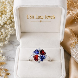 Könnte beinhalten: Ein silberfarbener Ring mit vier herzförmigen Edelsteinen in abwechselnd roten und blauen Farben, akzentuiert durch kleine klare Steine, präsentiert in einer weißen Samtschmuckschatulle mit dem goldenen Schriftzug USA Luxe Jewels im Deckel.