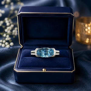 May include: A rectangular aquamarine ring set in a white metal band, presented in a navy blue jewellery box. The ring is surrounded by smaller, clear gemstones. The box is lined with blue velvet and rests on a dark blue satin surface.