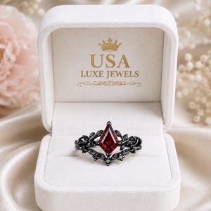 May include: A black metal ring featuring a kite-shaped red gemstone center stone surrounded by small black stones and leaf-like metal details. The ring is displayed inside a white velvet box with the text USA LUXE JEWELS and a crown icon printed.