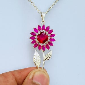 Puede incluir: Un collar con colgante plateado con un diseño floral. La flor tiene un gran centro de piedra preciosa roja redonda rodeado de piedras preciosas rosas más pequeñas. El colgante incluye dos acentos de hojas y cuelga de una delicada cadena.