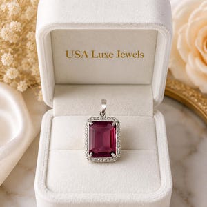 May include: A rectangular pendant featuring a large, deep magenta gemstone set in a silver-toned frame surrounded by small clear stones. The pendant is displayed inside a white velvet jewelry box with the text USA Luxe Jewels printed in gold.