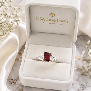 May include: A silver-toned ring featuring a rectangular red center stone flanked by three smaller clear stones on each side. The ring is displayed inside a cream-colored velvet jewelry box with the text USA Luxe Jewels printed in gold on the lid.