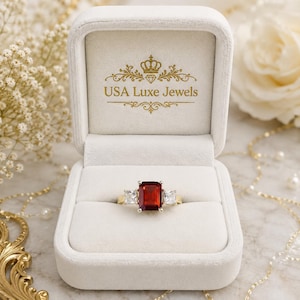 Puede incluir: Un anillo de oro con una gran piedra roja de corte esmeralda flanqueada por dos piedras claras de corte cuadrado, presentado en un estuche de terciopelo blanco con el texto dorado USA Luxe Jewels y un icono de corona.