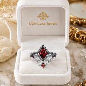 May include: A silver ring featuring a central coffin-shaped red gemstone, accented with smaller black stones and a decorative filigree band. The ring is displayed inside a white velvet jewelry box with the gold text USA Luxe Jewels printed inside.