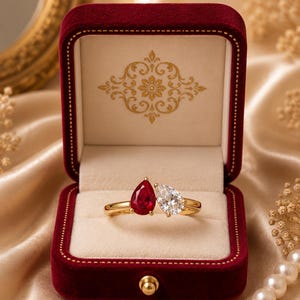 Può includere: Un anello d'oro con due gemme a forma di pera, una rossa e una trasparente, presentato in un cofanetto di velluto rosso scuro. Il cofanetto ha una chiusura dorata e si trova su un tessuto dorato morbido.