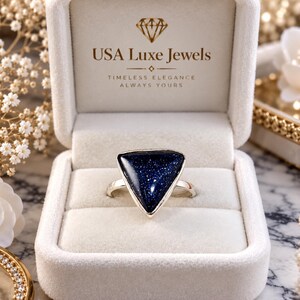 Pode incluir: Um anel de prata com uma pedra de arenito azul escuro triangular com inclusões brilhantes, apresentado numa caixa de joias de veludo creme com o texto USA Luxe Jewels, Timeless Elegance, Always Yours na tampa interior.