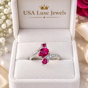 May include: A silver ring featuring a large oval-cut red ruby centre stone flanked by two smaller round-cut red rubies and accented with small clear stones along the band, presented in a white box labelled with the text USA Luxe Jewels.