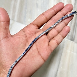 May include: A silver-toned tennis bracelet featuring a continuous row of square-cut blue gemstones set in a metallic channel. The bracelet displays a uniform design with a secure clasp mechanism at one end, showcasing a sleek and polished appearance.