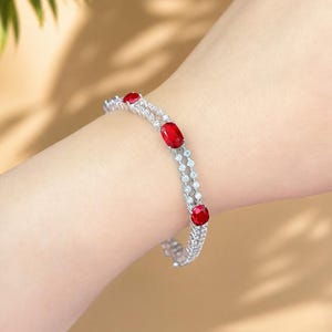 May include: A silver bracelet featuring three oval-cut red gemstones and many smaller clear stones. The bracelet is displayed on a wrist, highlighting the design and sparkle. The bracelet is a piece of fine jewellery.