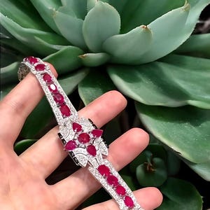 May include: A silver bracelet featuring oval-cut red gemstones and small, clear accent stones. The bracelet has a floral design in the centre and is set against a backdrop of green succulent leaves.