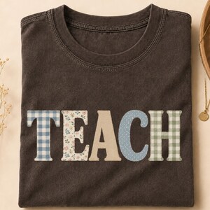 Puede incluir: Una camiseta de color marrón oscuro con la palabra TEACH impresa en letras grandes de bloque en el pecho. Cada letra tiene un patrón único: cuadros vichy azules, estampado floral, beige liso, azul con puntos blancos y cuadros vichy verdes.