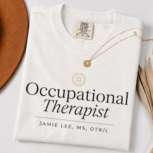 Puede incluir: Una camiseta blanca de manga corta con un gráfico de botón dorado sobre el texto negro Occupational Therapist y el nombre JAMIE LEE, MS, OTR/L. La camiseta tiene una etiqueta de la marca Comfort Colors en el cuello y un collar dorado.