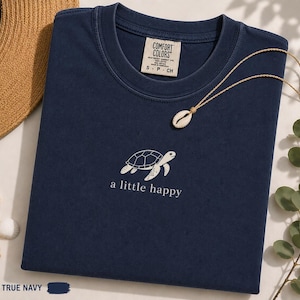 Puede incluir: Una camiseta azul marino con un gráfico bordado de una tortuga marina blanca en el centro del pecho y el texto a little happy impreso debajo. La etiqueta de la marca Comfort Colors en el cuello interior indica la talla S, P, CH.