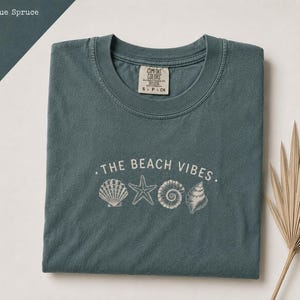 Puede incluir: Una camiseta de color azul abeto con un estampado gráfico blanco de cuatro conchas y una estrella de mar bajo el texto THE BEACH VIBES. La camiseta tiene cuello redondo y una etiqueta de la marca Comfort Colors en el interior del cuello.