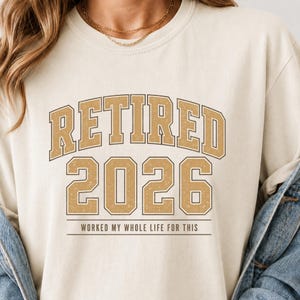 Puede incluir: Una sudadera de cuello redondo color crema con el texto RETIRED 2026 en una fuente universitaria dorada desgastada. Debajo del año, la frase WORKED MY WHOLE LIFE FOR THIS está impresa en una tipografía sans-serif pequeña y sencilla.