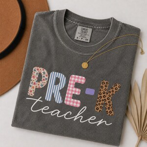 Puede incluir: Una camiseta de manga corta gris carbón con cuello redondo. La parte delantera presenta el texto "PRE-K teacher" en una mezcla de estampados florales, rayas azules, cuadros vichy rosas y leopardo. Un collar de cadena dorada descansa sobre la tela.