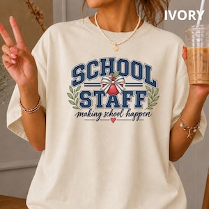 May include: An ivory t-shirt featuring a graphic with the text "SCHOOL STAFF" in dark blue block letters. Between the words is a red apple illustration with a striped bow. Below, the text reads "making school happen" with a small red heart.