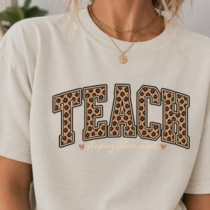 Puede incluir: Una camiseta de manga corta de color crema con la palabra TEACH en letras grandes de estilo bloque con un estampado de leopardo marrón y negro. Debajo de la palabra, la frase shaping future minds está impresa en cursiva con corazones.