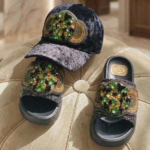 Puede incluir: Una gorra de béisbol de terciopelo negro y un par de sandalias negras a juego. Ambos artículos presentan un adorno circular grande en la parte delantera, decorado con grupos de cristales verdes y ámbar rodeados de abalorios.