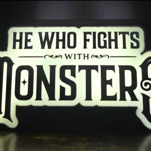 May include: A rectangular, illuminated sign with a cream-colored background and black lettering. The text reads HE WHO FIGHTS WITH MONSTERS in a stylized, bold font. The sign features decorative flourishes and a star-like accent within the letter O.