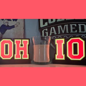 May include: Two separate rectangular wooden signs featuring the letters O and H on the left and I and O on the right, all in bright red with yellow outlines. A clear glass candle holder containing a light-colored wax candle sits between them.