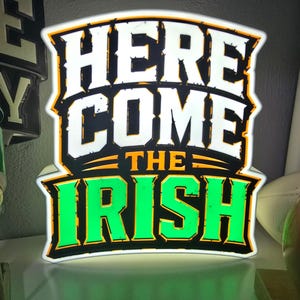 May include: A rectangular illuminated sign featuring the text "HERE COME THE IRISH" in bold, stylized lettering. The words "HERE COME" are in white, while "THE IRISH" is in bright green, all outlined in black and yellow against a glowing background.