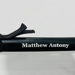 May include: A black sword scabbard featuring a smooth, glossy finish and a black fabric cord wrapped around the top section. The name Matthew Antony is printed in white, serif lettering on the side of the scabbard, centred horizontally.