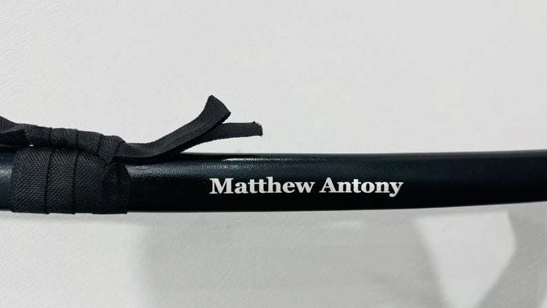 May include: A black sword scabbard featuring a smooth, glossy finish and a black fabric cord wrapped around the top section. The name Matthew Antony is printed in white, serif lettering on the side of the scabbard, centred horizontally.