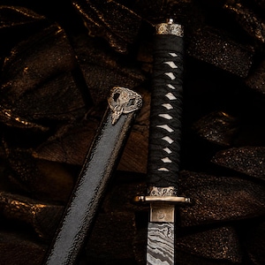 May include: A short sword with a patterned Damascus steel blade, a black cord-wrapped handle, and an ornate bronze-coloured guard and pommel. The matching black scabbard features a detailed bronze-coloured emblem at the top with a winged design.
