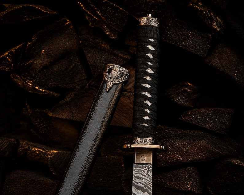May include: A short sword with a patterned Damascus steel blade, a black cord-wrapped handle, and an ornate bronze-coloured guard and pommel. The matching black scabbard features a detailed bronze-coloured emblem at the top with a winged design.