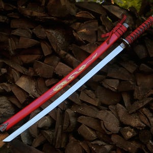 May include: A katana sword with a silver blade and a red scabbard featuring a gold dragon design. The handle is wrapped in red and black cord with a silver pommel. A red cord is tied near the top of the scabbard, which has a silver metal tip.