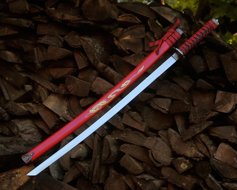May include: A katana sword with a silver blade and a red scabbard featuring a gold dragon design. The handle is wrapped in red and black cord with a silver pommel. A red cord is tied near the top of the scabbard, which has a silver metal tip.