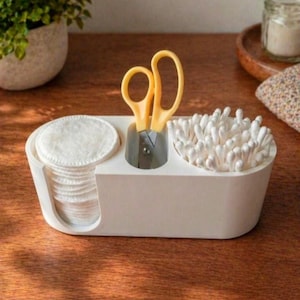 May include: A white oval-shaped vanity organiser featuring three distinct compartments. The left section holds a stack of round white cotton pads, the centre holds a pair of yellow-handled scissors, and the right section contains white cotton swabs.