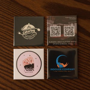Op de afbeelding: Vier vierkante magneten met logo's. Eén zwarte magneet heeft een cateringlogo met de tekst "Best Street Food". Een andere heeft een website en telefoonnummer met QR-codes. Een derde magneet heeft een nagelsalonlogo. De laatste magneet heeft een logo van een adviesbureau.