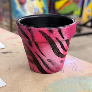 May include: A small, empty flower pot with a pink and black zebra stripe pattern. The interior of the pot is black. The pot is sitting on a light brown surface.
