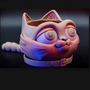 May include: A matte, light-toned ceramic planter shaped like a cat head with large, wide-set eyes, pointed ears, and a segmented tail extending from the side. The base features a sculpted collar detail with a buckle, creating a rounded vessel.
