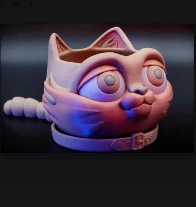 May include: A matte, light-toned ceramic planter shaped like a cat head with large, wide-set eyes, pointed ears, and a segmented tail extending from the side. The base features a sculpted collar detail with a buckle, creating a rounded vessel.