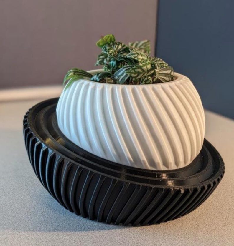 May include: A white, spherical planter with a diagonal ribbed texture sits inside a black, bowl-shaped base with vertical ridges. A small green plant with variegated leaves is planted inside the white container, which rests at an angle.