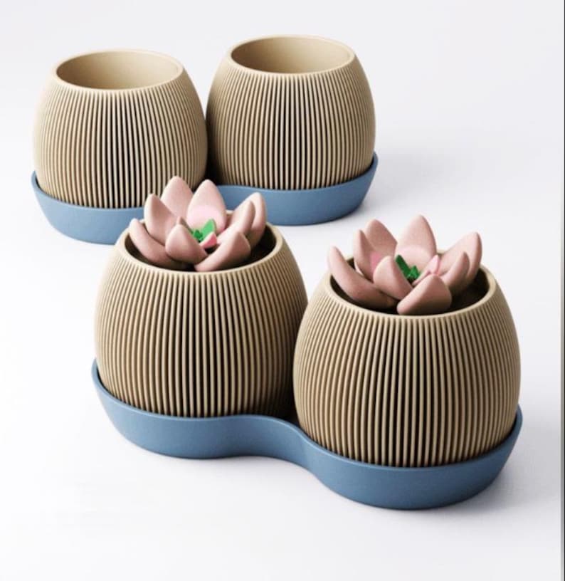 May include: Four round, ribbed beige planters with blue saucers. Two planters are empty, and two contain artificial pink succulent plants with green centres. The planters feature a vertical line texture and sit on matching blue bases.