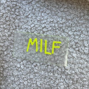 Könnte beinhalten: Ein rechteckiger, durchscheinender Aufkleber mit dem Text MILF in leuchtend neongelber Tinte. Der Aufkleber befindet sich auf einer strukturierten, cremefarbenen Stoffoberfläche neben einem beigen Stoffstück und einem getrockneten Pflanzenstängel.