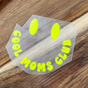 Könnte beinhalten: Ein transparenter Aufkleber mit einem leuchtend neongelben Smiley-Gesicht. Der Schriftzug COOL MOMS CLUB ist in einer fetten, abgerundeten Schrift entlang der unteren Gesichtskurve gedruckt, mit zwei vertikalen ovalen Augen über dem Text.