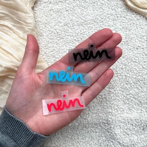 Könnte beinhalten: Drei rechteckige, halbtransparente Klebeetiketten, die jeweils das Wort "nein" in Schreibschrift zeigen. Das oberste Etikett ist schwarz, das mittlere hellblau und das untere rot, jeweils mit einem kleinen Punkt über dem Wort.