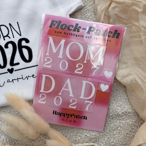 Könnte beinhalten: Ein weißer Babybody mit schwarzem Text 'RN 026 arrive' und eine rosa rechteckige Verpackung für Aufbügler mit der Aufschrift 'Flock-Patch zum Aufbügeln auf Textilien', weißem Text 'MOM 2027' und 'DAD 2027' sowie kleinen weißen Herzsymbolen.