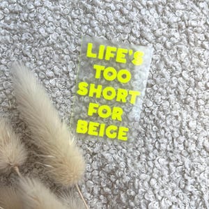 Könnte beinhalten: Ein rechteckiger, durchsichtiger Aufkleber mit leuchtend neongelber Schrift, die LIFE'S TOO SHORT FOR BEIGE lautet, platziert neben drei getrockneten beigen Ziergräsern mit weichen, federartigen Texturen auf einem lockigen, cremeweißen Stoffhintergrund.