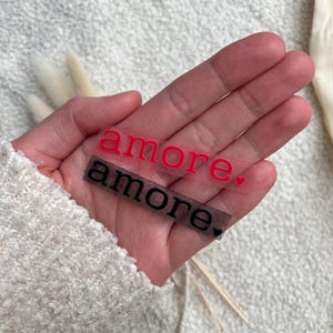 Könnte beinhalten: Zwei transparente Aufkleber mit dem Wort "amore" in Rot und Schwarz. Der rote Aufkleber hat ein kleines Herz. Die Aufkleber werden in einer Hand vor einem strukturierten weißen Hintergrund gehalten.