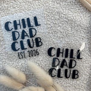 Könnte beinhalten: Zwei transparente Wärmeübertragungsfolien mit dem Text "CHILL DAD CLUB" in Schwarz. Eine Folie enthält auch "EST. 2026". Die Folien befinden sich auf einer strukturierten, cremefarbenen Oberfläche, mit getrocknetem Hasenschwanzgras und einem cremefarbenen Stoff im Hintergrund.